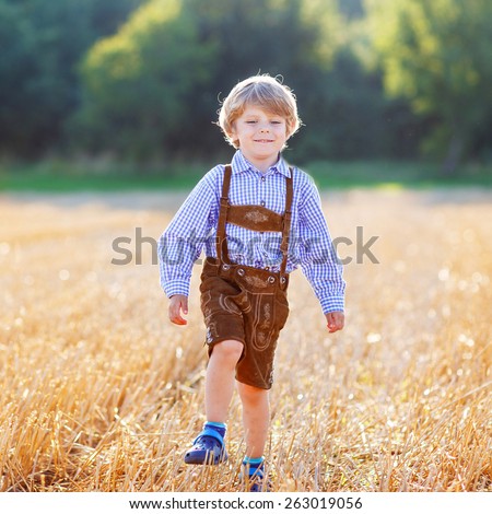 Funny little kid boy in traditional German bavarian clothes, leather shorts and check shirt, walking happily through wheat field near  hay stack or bale. Active outdoors leisure with children