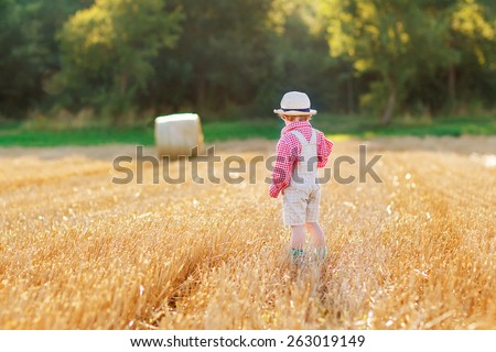 Funny little toddler boy in traditional German bavarian clothes, leather shorts and check shirt,  walking happily through wheat field near  hay stack or bale.