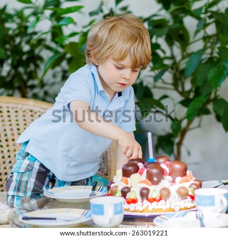 Funny blond boy celebrating his third birthday in home\'s garden with big cake. Happy child laughing about gifts and tasting cake. Child helping and serving table