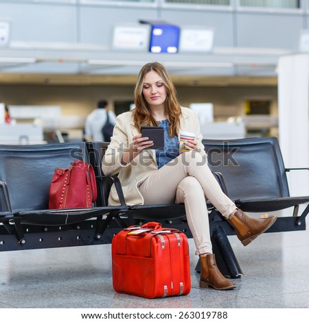 Young woman at international airport, reading her ebook computer and drinking coffee to go while waiting for her flight. Female passenger at terminal, indoors.