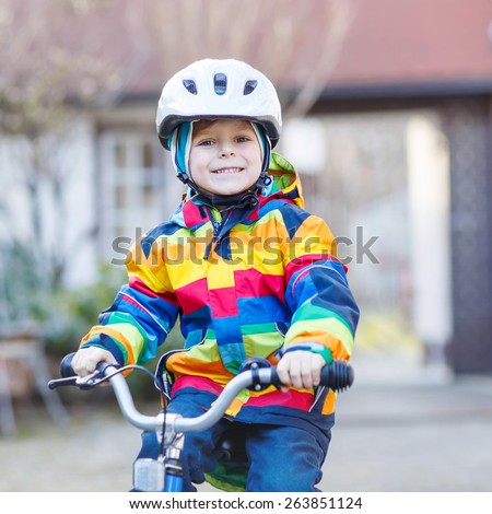 Funny cute  preschool kid boy in safety helmet and colorful raincoat riding his first bike and having fun on cold  day, outdoors. Active leisure with children in winter, sping or autumn.