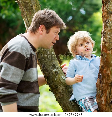 Father and his little cute son having fun in forest. Funny kid boy climbing on tree. Family leisure outdoors, on warm summer day.