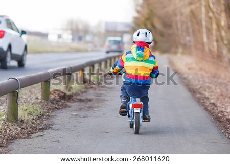 Little kid boy in safety helmet and colorful raincoat riding his first bike and having fun on cold  day, outdoors. From back, street with cars.