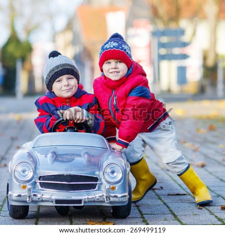 Two happy sibling boys in red jackets and rain boots playing with big old toy car, outdoors.  Kids leisure on cold day in winter, autumn or spring.