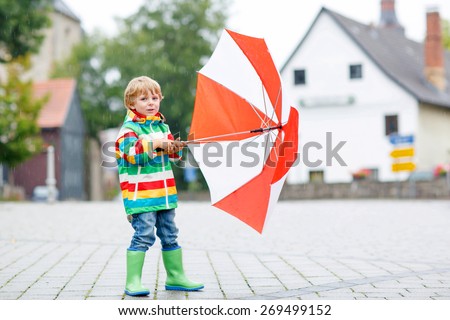 Preschool boy in waterproof clothes and boots walking on the street of a small town on rainy day.