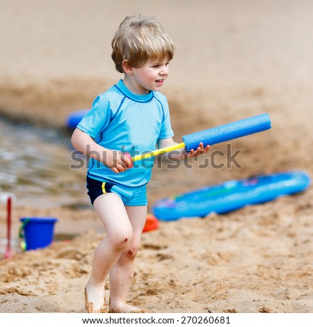Little blond child having fun with splashing water near city lake, playing water gun. Outdoors leisure with kids in summer, on sunny hot day.