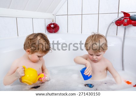 Twins having fun with water by taking bath in bathtub at home. Kids playing and cooperating together with toys. Children cleaning and washing, healthcare concept.