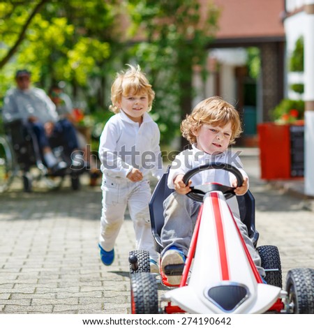 Two happy sibling boys playing with toy car in summer garden, outdoors. Children having fun in domestic garden or nursery on sunny warm day. With grandfather in wheelchair on background