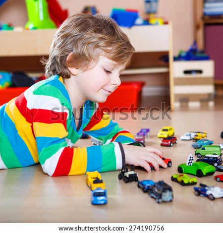 Happy funny little blond child playing with lots of toy cars indoor. Kid boy wearing colorful shirt and having fun at nursery.
