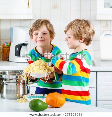 Two funny kid boys of 4 years cooking pasta and fresh vegetables in domestic kitchen, indoors. Sibling children in colorful shirts.