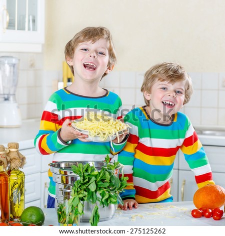 Two funny twin kids boys cooking and eating meal with spaghetti and fresh vegetables in domestic kitchen, indoors. Sibling children in colorful shirts.