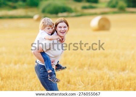 Adorable blond kid boy and his mother having fun on yellow hay field in summer. Happy family of two enjoying nature and togetherness. Boy riding on mum.