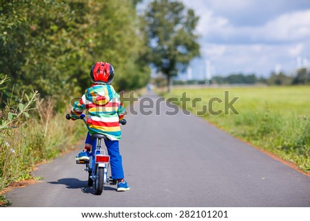 Little child in red safety helmet and colorful raincoat riding his first bike on summer day. Active leisure for kids outdoors.
