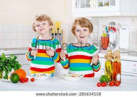 Two funny twin kids boys cooking and eating meal with spaghetti and fresh vegetables in domestic kitchen, indoors. Sibling children in colorful shirts.