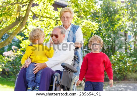 Two little kid boys, their grandmother and grandfather in wheelchair in summer garden. Happy family spending time together.