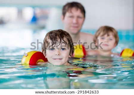 Young father teaching his two little sons to swim in an indoor swimming pool. Active happy kid boys, siblings and twins wearing safe swimmies and having fun together.