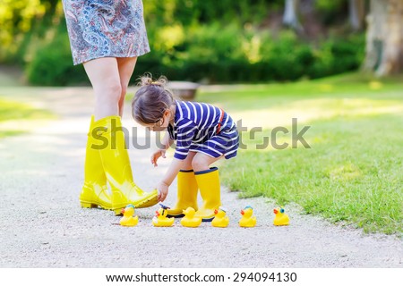 Woman and her little cute kid daughter in yellow rubber boots, family look, playing with duck toys in summer park. Active leisure with kids.