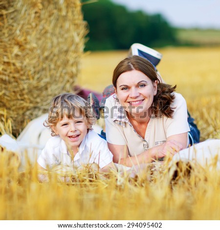 Young mother and her little son having fun at picnicking on yellow hay field in summer. Happy family of two enjoying nature and togetherness.
