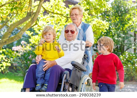 Sibling kids, their grandmother and grandfather in wheelchair in summer garden. Happy family spending time together.