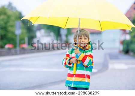 Little blond kid boy walking with big umbrella outdoors on rainy day. Child having fun and wearing colorful waterproof clothes and rain boots.