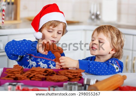 Two little kid boys in red santa hat with fresh baked gingerbread cookies. Happy about gingerbread man. Kitchen decorated for Christmas