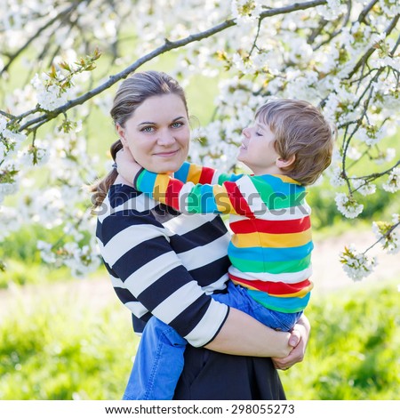 Young woman and her little son, blond child hugging in blooming cherry garden in spring. Happy family celebrating mother\'s day.