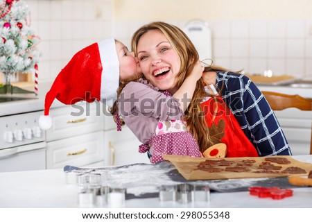Happy mother, young woman in red santa hat baking christmas cookies together with her little daughter, cute blonde kid girl, sitting at the table in domestic kitchen