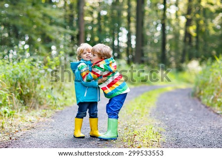 Two little kid boys, brothers in colorful waterproof raincoats and rubber boots hugging in autumn forest together.