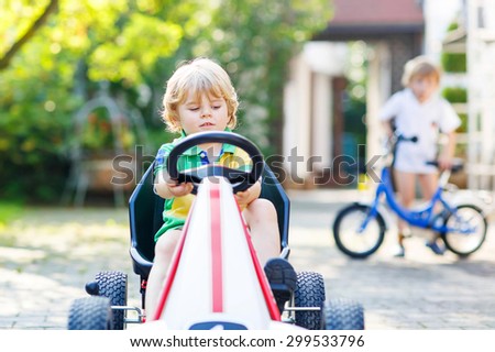Active little boy of 3 years driving pedal car in summer garden, outdoors. His little brother on bike on background. On warm sunny day.