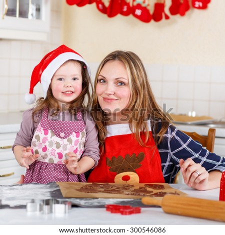 Happy mother, young woman in red santa hat baking christmas cookies together with her little daughter, cute blonde kid girl, sitting at the table in domestic kitchen