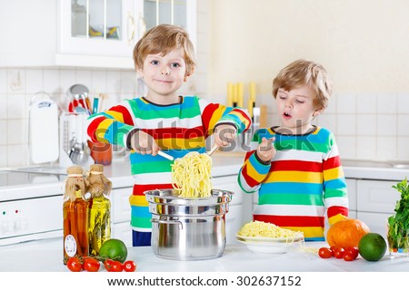 Two little friends having fun with cooking of spaghetti and fresh vegetables in domestic kitchen, indoors. Sibling children in colorful shirts.