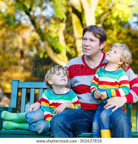 Young man and his two little blond sons sitting together in colorful clothing. Happy kid boys and their dad having fun in autumn park on warm day.