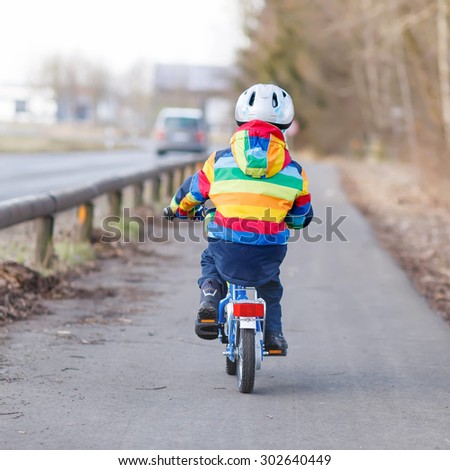 Little kid boy in safety helmet and colorful raincoat riding his first bike and having fun on cold  day, outdoors. From back, street with cars.