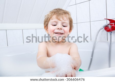 Funny blond kid boy having fun with water by taking bath in bathtub at home. Lovely child splashing and playing with foam.