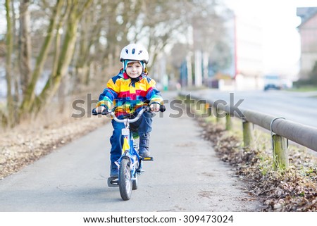 Happy boy of 5 years in safety helmet and colorful raincoat riding his first bike and having fun on cold  day, outdoors. Active leisure with children in winter, spring or autumn.