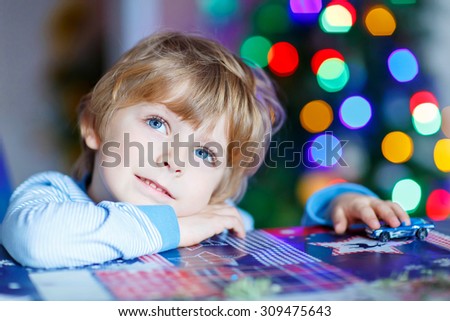 Little blond child playing with cars and toys at home, indoor. Cute happy funny boy having fun with gifts. Colorful christmas lights on background. Family, holiday, kids lifestyle concept.