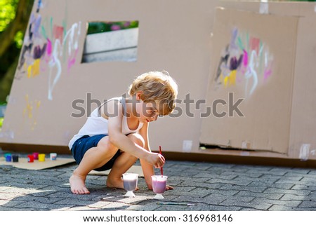 Adorable little kid boy painting big paper house with colorful paintbox. Children having fun outdoors. Creative leisure, preschool project for pupil.