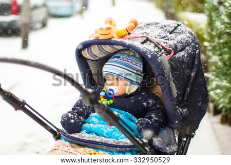 Cute baby boy in warm clothes in pram during winter snow fall on cold winter day. Happy carefree childhood.