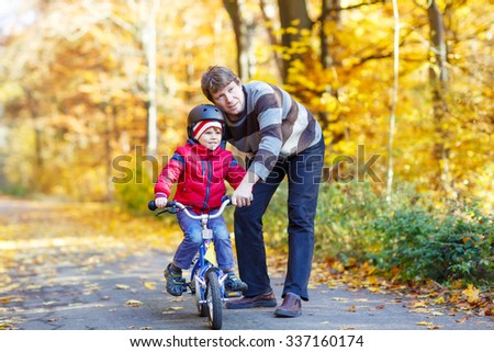 Young man and his little son, kid boy in autumn forest with a bicycle. Father teaching biking. Active family leisure. Child with helmet. Safety, sports, leisure with kids concept.