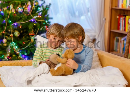 Two adorable little blond kid boys, twins playing with soft toys together in bed near Christmas tree with lights and illumination. Happy family, two children and friends.