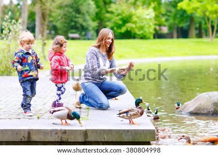 Mother and her children feeding ducks in summer park, adorable kid boy and girl, siblings having fun together