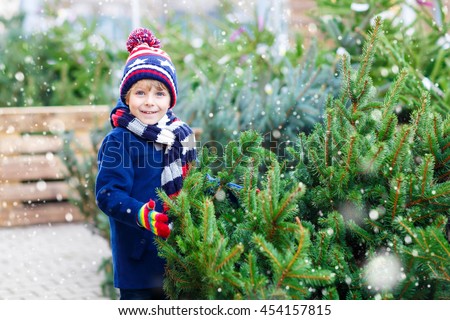 Adorable little smiling kid boy holding christmas tree. Happy child in winter clothes, hat, gloves choosing xmas tree in outdoor shop. Family, tradition, celebration concept