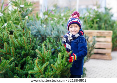 Funny little smiling kid boy holding christmas tree. Happy child in winter fashion clothes choosing and buying xmas tree in outdoor shop. Family, tradition, celebration