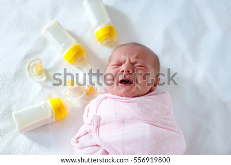 Crying newborn baby girl with nursing bottles. Formula drink for babies. New born child, little girl laying in bed. Family, new life, childhood, beginning, bottle-feeding concept.