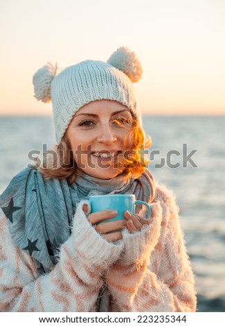 Cute young european woman in knitted clothes drinks coffee on the beach at sunset. Cozy, comfy, soft