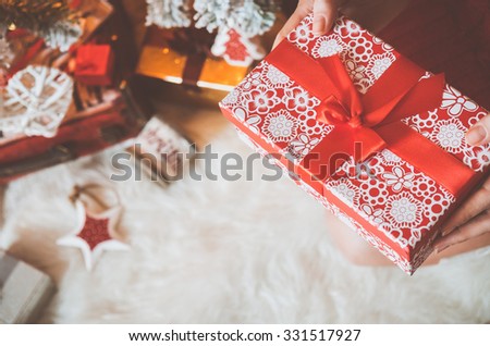 Young pretty woman in warm cozy clothes is sitting on the floor in her bright house near the christmas tree and holds gift box in her hands.