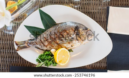 grilled dorado fish on a white plate with lemon and herbs on a glass table in a restaurant with a glass of white wine