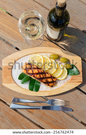 delicious salmon fillet grilled served on a wooden plate with lemon, olives and sea salt on wooden table in restaurant with white wine