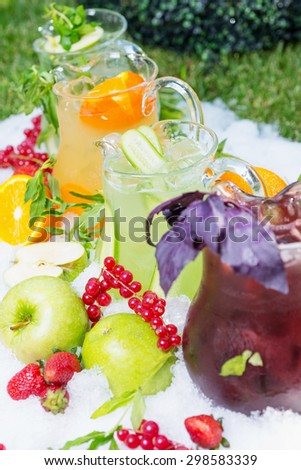 a mix of several bright colored delicious cold lemonade carafes of cucumber and orange and basilon with creative composition of ice berries oranges apples and mint. soft focus