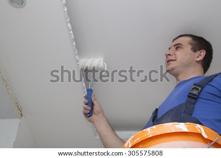 Man repairs the house inside and paints the ceiling holding roller and bucket with paint